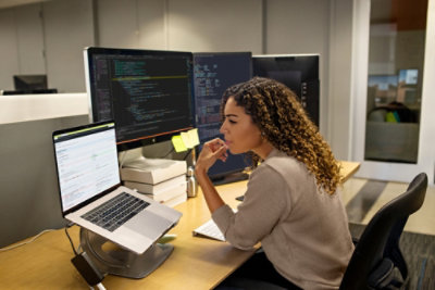 Egy nő ül egy asztalnál több képernyővel, és egy laptopot néz