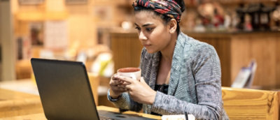 A woman with a head scarf holding a cup and looking at a laptop.