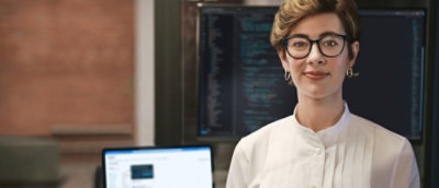 Picture of a woman wearing glasses with a laptop and a screen behind.