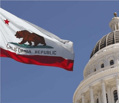 The California Republic flag waving in the foreground with the domed state capitol building visible against a clear blue sky.