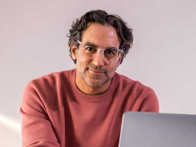 Un hombre sentado en un escritorio con un portátil con gafas y una camiseta roja; Texto verde y negro sobre un fondo blanco: ahorre hasta un 76 % en comparación con los precios de pago por uso.