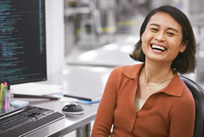 En smilende person, der sidder ved et skrivebord med en computer, der viser kodelinjer på skærmen.