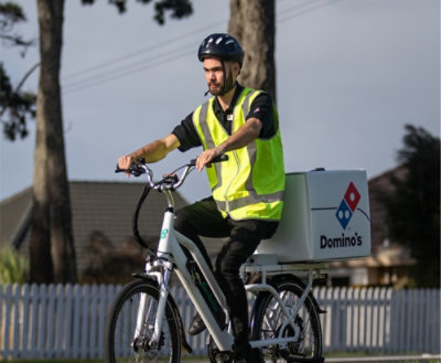 Pessoa andando de bicicleta com uma caixa de entrega de pizza, usando colete de segurança e capacete.