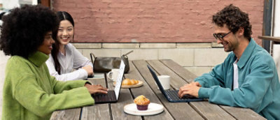  Three people working on laptops at an outdoor wooden table with coffee cups and pastries.