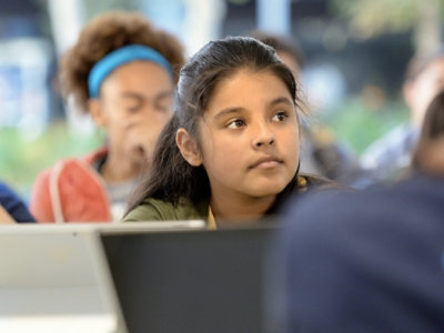 A young girl with long dark hair sits attentively in a classroom, looking slightly to the side.