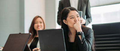 Une jeune femme asiatique en tailleur professionnel écoute intensément lors d’une réunion, avec un ordinateur portable ouvert, et un autre participant 