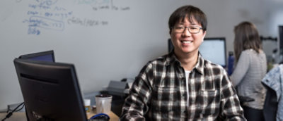 Une personne d’Asie de l’Est souriante, portant des lunettes et une chemise écossaise, est assise devant un ordinateur dans un bureau avec un tableau blanc 