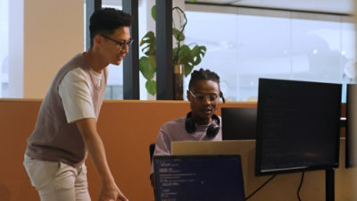 Two people in an office setting collaborating at a workstation. One person is standing and pointing toward a large monitor, while the other is seated at a desk with two screens displaying code. 