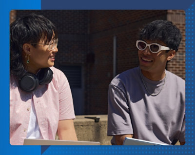 Zwei Personen lächeln sich draußen vor einem Backsteingebäude an einem sonnigen Tag an. Eine trägt Kopfhörer und eine Brille, die andere Sonnenbrille.
