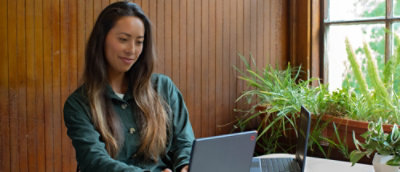 Woman smiling while using a laptop in a room with plants and wooden paneling.