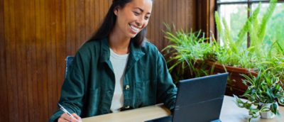 Egy íróasztalnál dolgozó személy laptopjával és jegyzetfüzetével egy növényekkel teli munkakörnyezetben