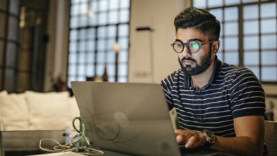 Personne travaillant sur un ordinateur portable à une table dans un espace intérieur faiblement éclairé, avec des écouteurs et une tasse de café à proximité.