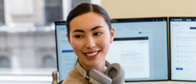 En kvinde med hovedtelefoner om halsen smiler til kameraet foran en computerskærm.