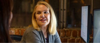 Blonde vrouw die achter een bureau zit en met een andere persoon praat. Er zijn twee computermonitors zichtbaar naast haar