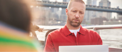 Een man met een baard en een rood overhemd is bezig op een laptop terwijl hij op een boot zit 