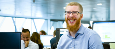 En man med rött skägg och glasögon som ler när han sitter vid sitt skrivbord i ett kontor med kollegor 