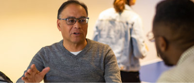 Mand med briller og grå sweater, der har en samtale med en anden person. En tredje person ses i baggrunden,