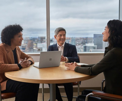 Three professionals, two women and a man, engaging in a discussion around a small table with a laptop