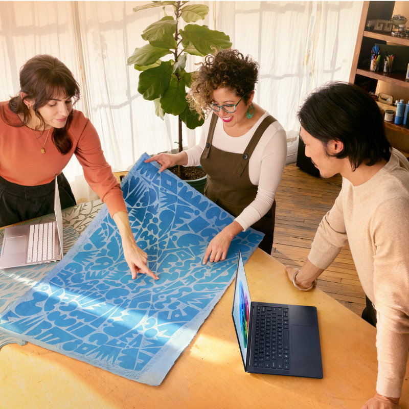 Drie mensen, in een open, lichte kamer met ramen met gordijnen, planten en een boekenplank op de achtergrond, leunen over een tafel met twee open laptops om naar een grote blauwe abstracte afdruk te kijken.