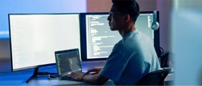 Seseorang bekerja di laptop dengan kode yang ditampilkan di dua monitor besar di ruangan yang redup.