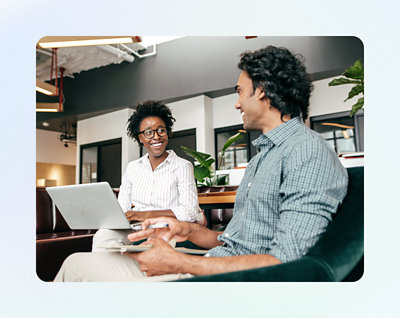 Two people are sitting in a modern office space, one holding a laptop and the other using a tablet, engaged in a conversation.