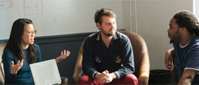 Tre personer sitter i en soffa och diskuterar med en whiteboard i bakgrunden.