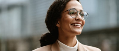 Une personne portant des lunettes et un manteau beige sourit en regardant au loin, avec un arrière-plan urbain flou.