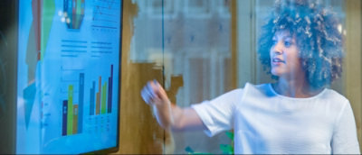 En person i hvit skjorte peker mot en skjerm som viser ulike stolpediagrammer og datavisualiseringer.