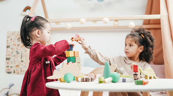 Two young girls playing with colorful wooden blocks and toys at a white table indoors.