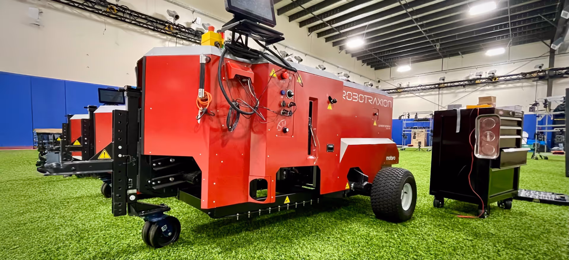 Red Robotraxion robot lawn mower on artificial turf in an indoor facility with tools nearby.
