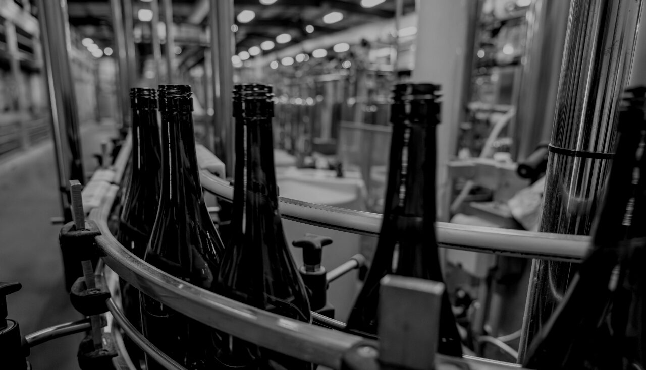 Dark glass bottles on a conveyor belt in a bottling or manufacturing facility with blurred industrial background.