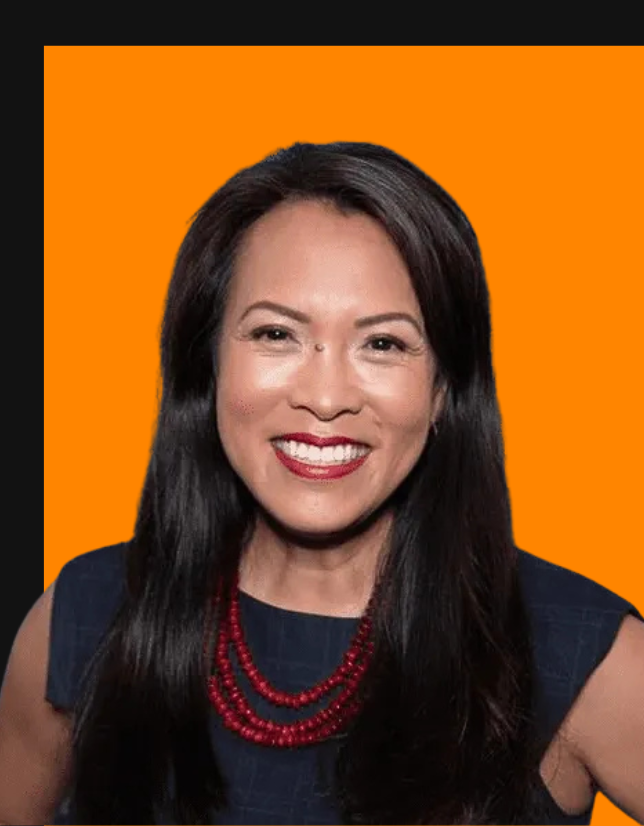 Smiling woman with long dark hair wearing a sleeveless dark top and red beaded necklace against an orange background.