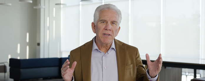 Mark McClain in a brown blazer speaking and gesturing in a bright office.
