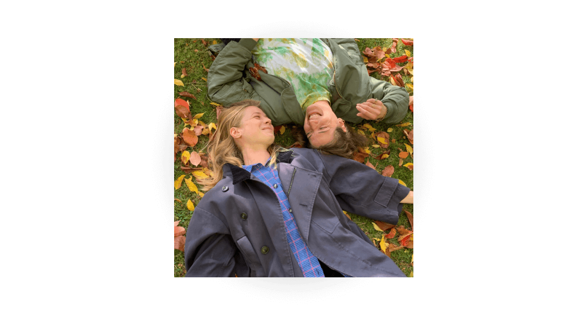 Two people lying on the grass side by side among fallen autumn leaves, smiling and looking toward each other.