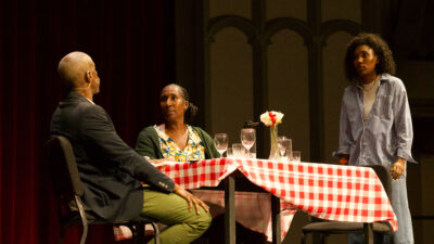 Three performers enact a tense family conversation around a table covered with a red-and-white checkered tablecloth.