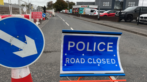 A photo on the Gortin Road, on the left is a circular blue sign with a white border, in the middle it has a white arrow pointing to another blue sign with a white border sign that reads 'Police road closed'.
In the background there is a Spar shop, a car park, and police barricades blocking the road.