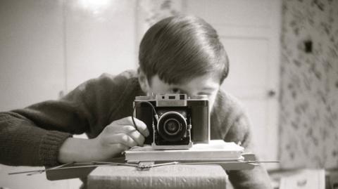 A child looks through the viewfinder of an old film camera in a grainy black and white image.
