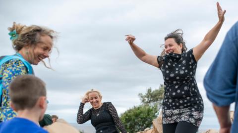 People jumping and walking around lots of cardboard. One woman in the centre of the photo is smiling and jumping up with her arms in the air.