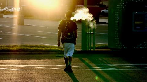 A man, Mike, walks across a road, as the sun sets, blowing a cloud of smoke into the air.