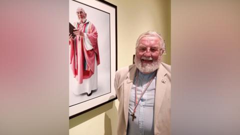 A man is standing next to a portrait of himself in robes as his job as a vicar. He is wearing a beige jacket, and a light blue shirt with a cross around his neck which is on a rainbow coloured lanyard. He has a white beard and is wearing glasses.
