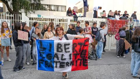 A high school student is holding a blue, white, and red tricolor flag bearing the inscription Liberte, Egalite, Fraternite in front of her high school.