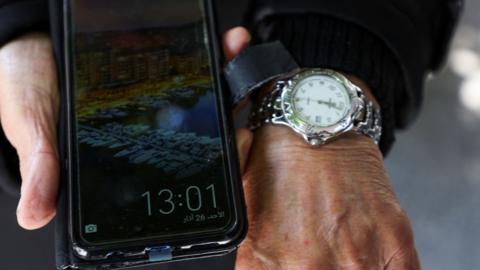 A smartphone in someones hands showing 13:01 and a watch on their wrist showing 12:01