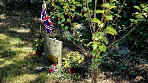 A gravestone next to an apple tree with a small Union Jack flag next to it. The stone reads Unknown, HMS Terror