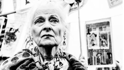 A black and white photo of Vivienne Westwood shows her looking into space as protest banners fly behind her.