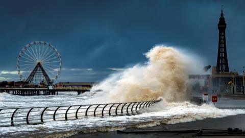 Large waves crash onto the promenade at Blackpool. Blackpool Tower and a ferris wheel are in the background.