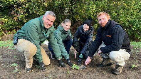 A group of four people leading the tower mustard conservation effort crouch down on a muddy ground with small shoots of plants in and smile at the camera. They are all touching one green plant.