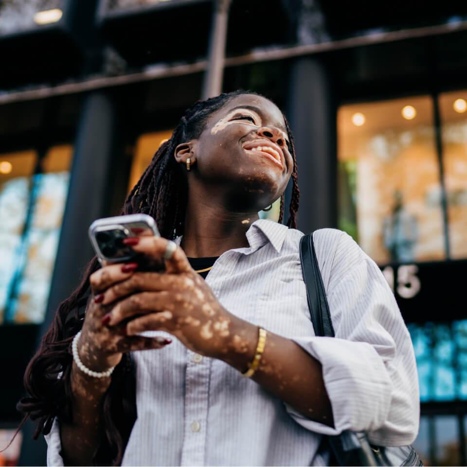 A smiling woman with vitiligo looking up happily while holding a smartphone in front of a modern building.