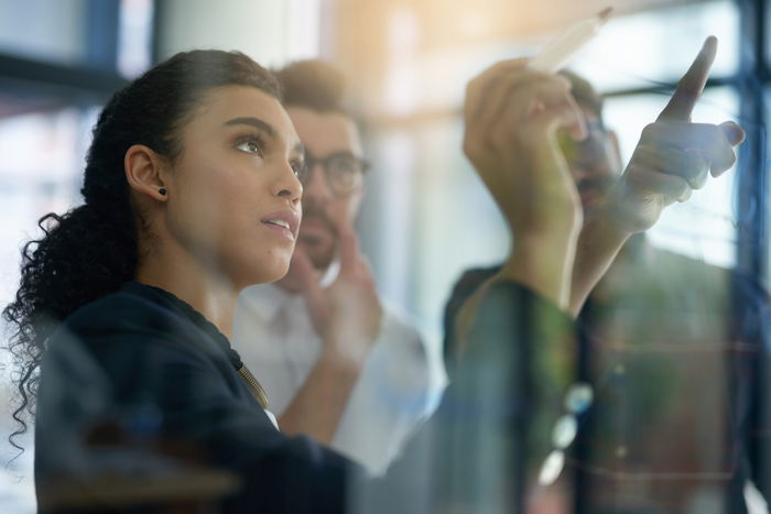 Three future talents are collaborating and brainstorming in front of a glass board, with one person writing or pointing at notes.