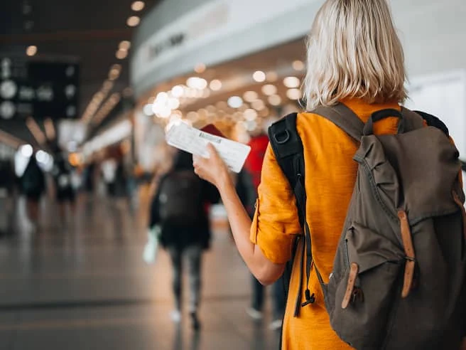 An individual standing in a crowded airport, wearing a backpack and holding a plane boarding pass.