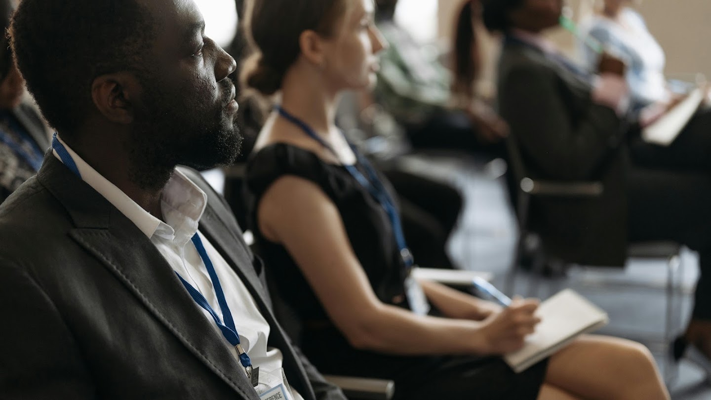 A side-profile view of attendees seated in an audience at a conference or seminar. In the sharp foreground on the left, a Black man with a beard, wearing a dark suit jacket over a white collared shirt and a blue conference lanyard, looks forward attentively. Seated next to him, slightly out of focus, a woman in a black sleeveless top with a blue lanyard holds a pen and rests a notepad on her lap. Other audience members are visible in the blurred background.